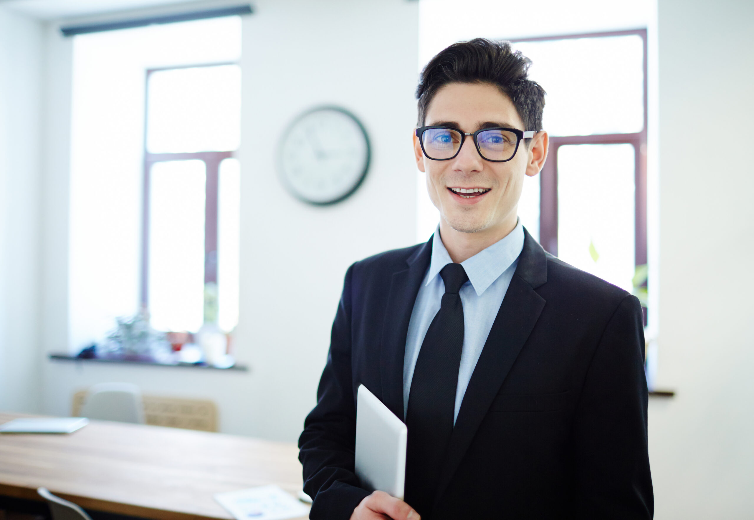 A smiling young professional holding a tablet, standing in a bright, modern office setting.