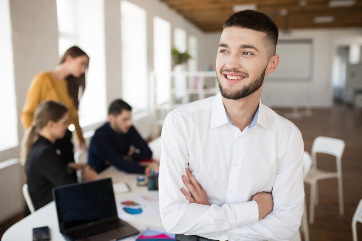 A confident young professional with his arms crossed, with his colleagues brainstorming in the background.